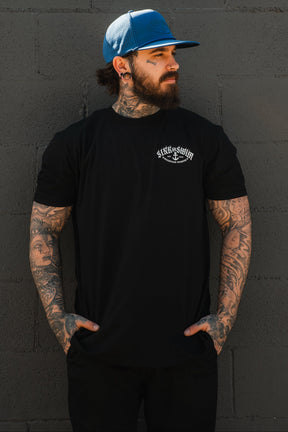 A bearded, tattooed man stands against a gray brick wall, wearing a Relentless Betrayal Dark Waters SOS T-Shirt, black pants, and a blue baseball cap. He looks to the side with his hands in his pockets.