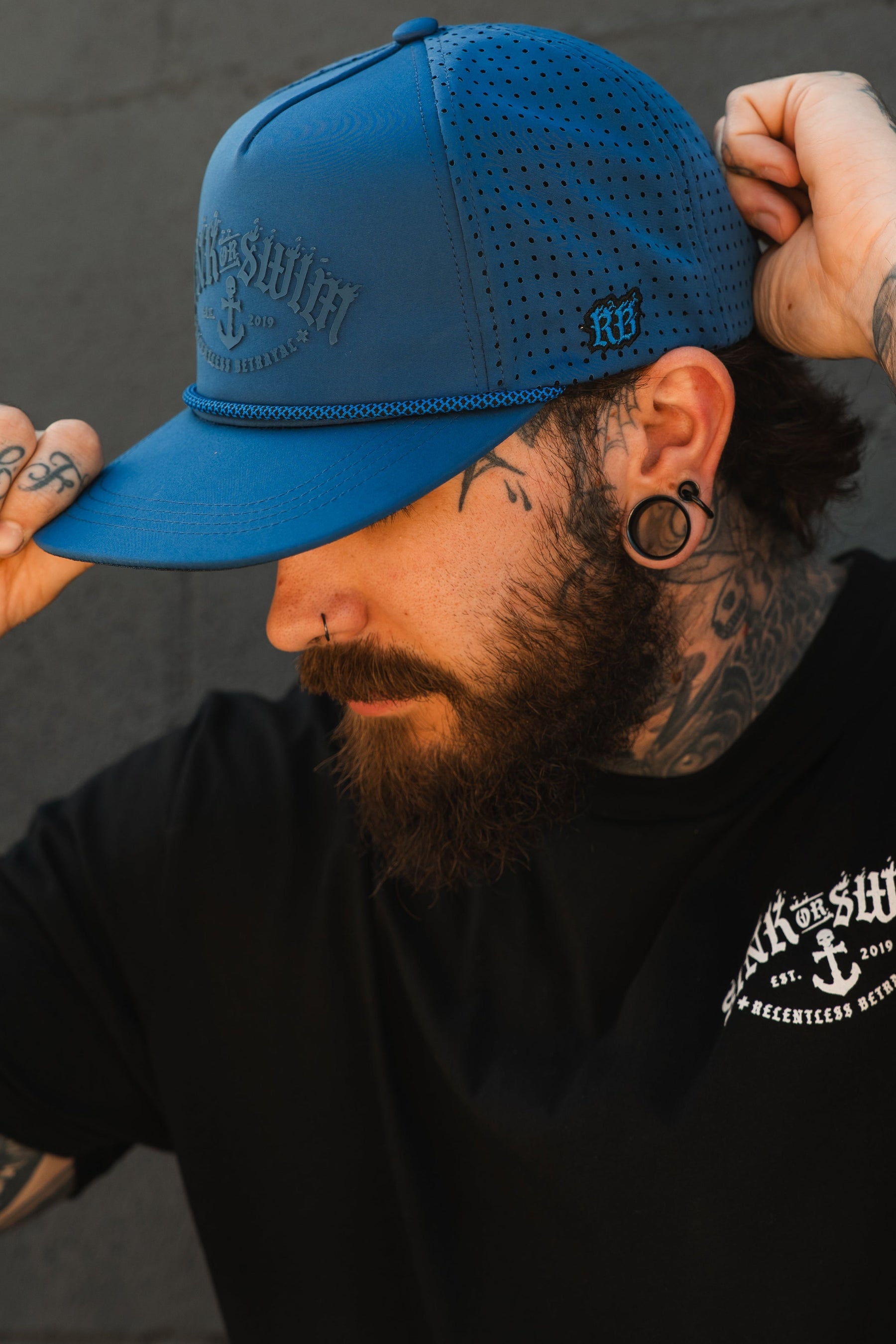 A tattooed, bearded person with stretched earlobes adjusts the Relentless Betrayal Dark Waters SOS Mute PVC Snapback, a black water-resistant hat with a blue rubber patch, while wearing a black T-shirt with white text against a dark background.