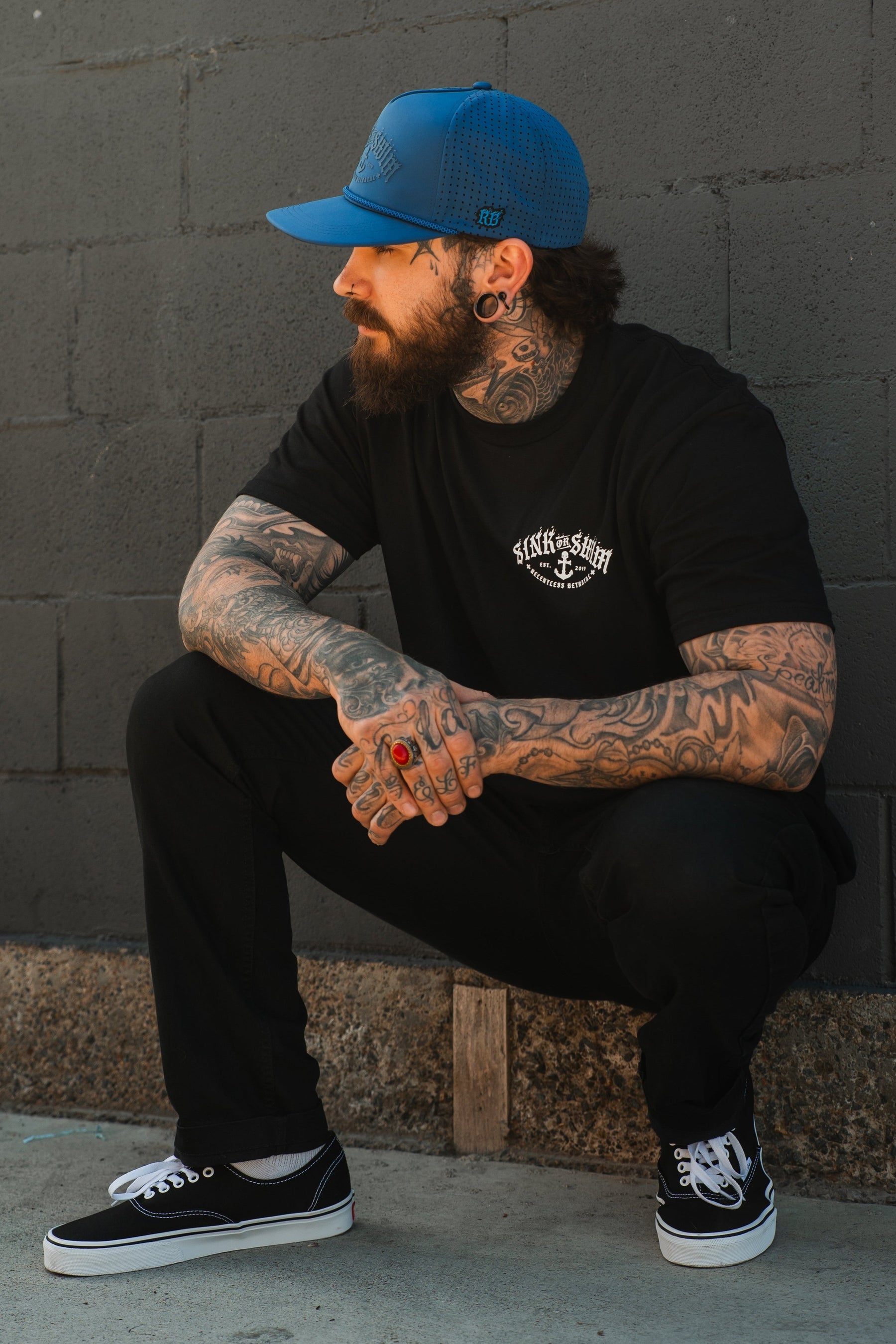 A bearded, tattooed man in a blue cap, black pants, and black Vans sneakers squats by a gray brick wall, looking to the side while wearing the Relentless Betrayal Dark Waters SOS T-Shirt—crafted from fine jersey cotton.