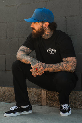 A bearded, tattooed man in a blue cap, black pants, and black Vans sneakers squats by a gray brick wall, looking to the side while wearing the Relentless Betrayal Dark Waters SOS T-Shirt—crafted from fine jersey cotton.