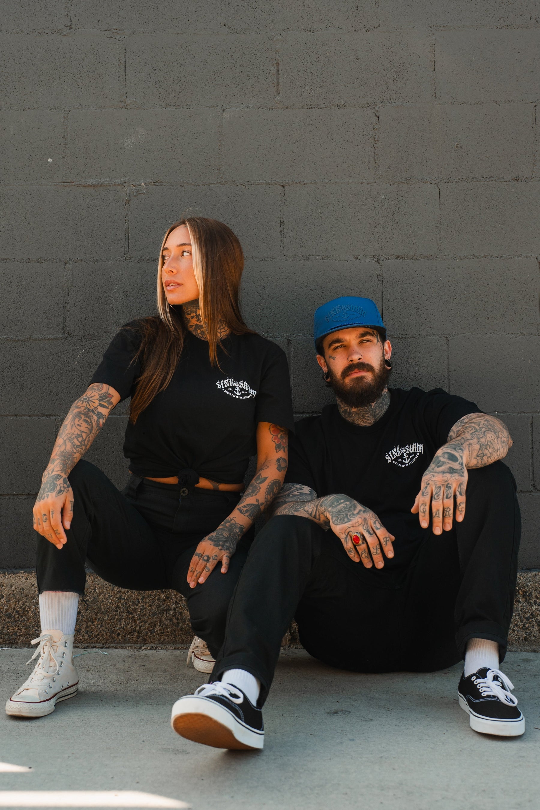 Two tattooed people in black unisex tees sit against a gray brick wall. The woman on the left wears white sneakers; the man on the right sports a blue cap, black Vans, and a Relentless Betrayal Dark Waters SOS T-Shirt in fine jersey cotton.