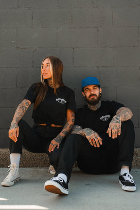 Two tattooed people in black unisex tees sit against a gray brick wall. The woman on the left wears white sneakers; the man on the right sports a blue cap, black Vans, and a Relentless Betrayal Dark Waters SOS T-Shirt in fine jersey cotton.