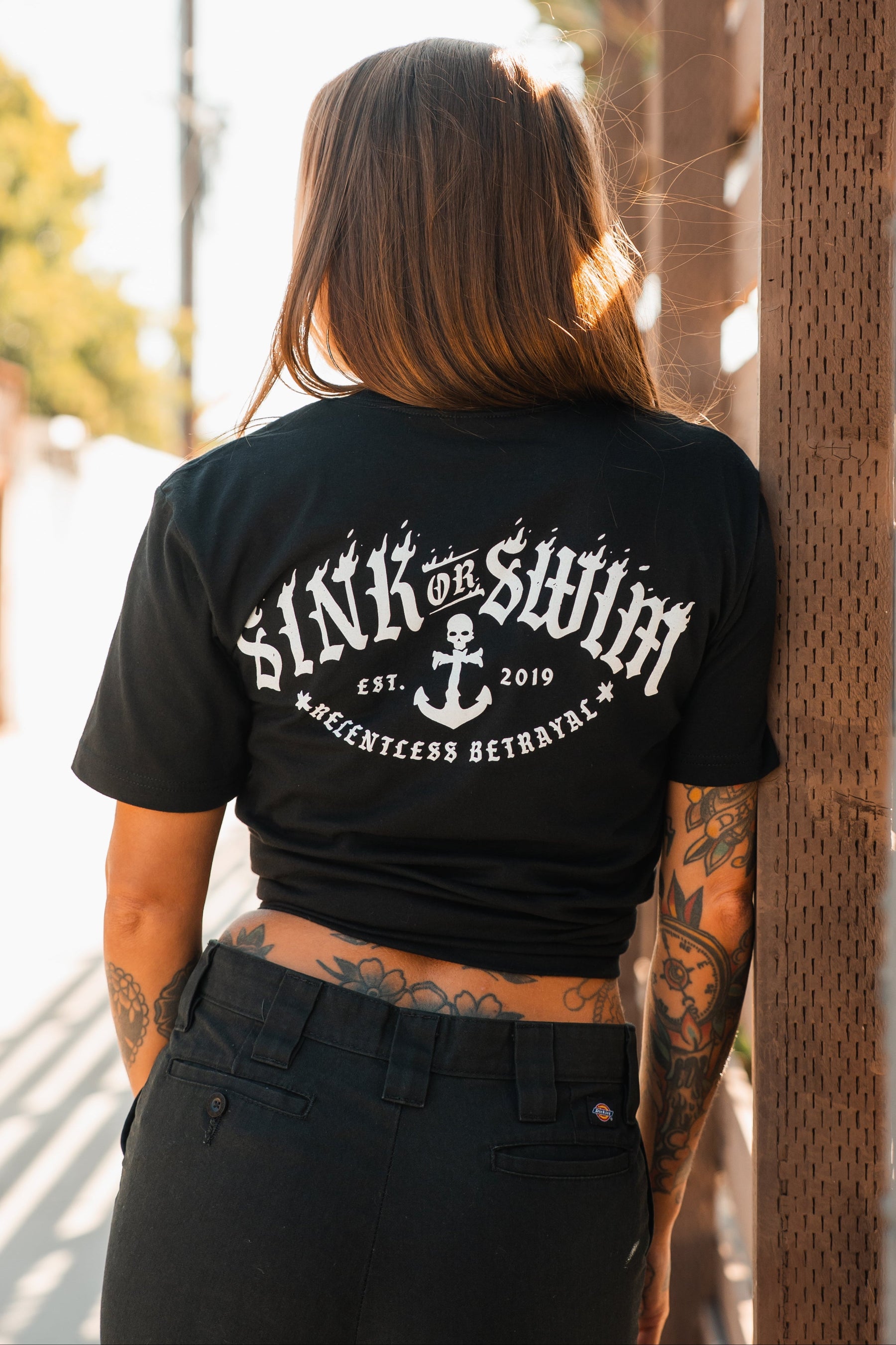 A tattooed woman stands outside, back turned, wearing the black Relentless Betrayal Dark Waters SOS T-Shirt featuring “SINK OR SWIM EST. 2019 RELENTLESS BETRAYAL” and an anchor graphic, paired with dark pants in the sunlight.