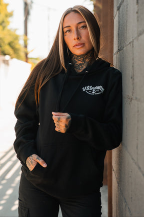 A woman with long brown hair, light highlights, and neck and hand tattoos wears a black Relentless Betrayal Dark Waters SOS Hoodie while leaning against a gray brick wall on a sunny day.