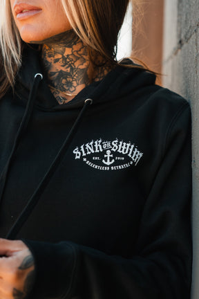 A woman with neck tattoos and blonde-streaked hair wears the Relentless Betrayal Dark Waters SOS Hoodie, a unisex cotton-poly hoodie with a white Sink or Swim anchor logo and "Relentless Betrayal Est. 2019" on the chest.