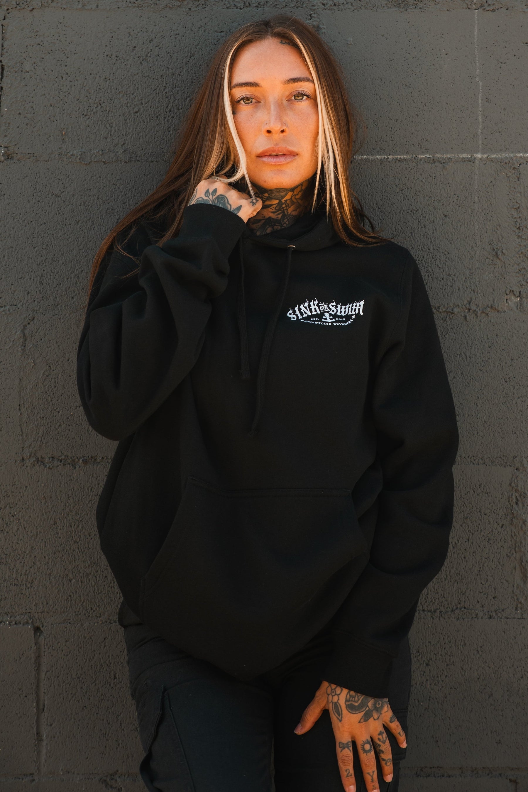 A person with long brown hair and facial tattoos wears a Relentless Betrayal Dark Waters SOS Hoodie—a unisex cotton-polyester hoodie with white lettering—while standing against a textured dark gray brick wall, looking slightly upward.