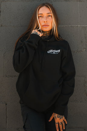 A person with long brown hair and facial tattoos wears a Relentless Betrayal Dark Waters SOS Hoodie—a unisex cotton-polyester hoodie with white lettering—while standing against a textured dark gray brick wall, looking slightly upward.