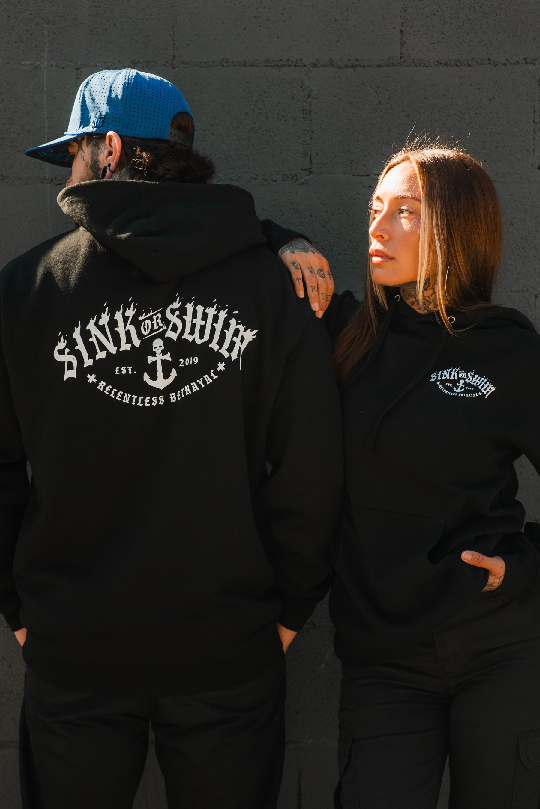 A man and woman wear Relentless Betrayal's Dark Waters SOS Hoodie by a gray brick wall; he shows off the back design, she faces him with her hand on her hip. This unisex hoodie elevates any look.