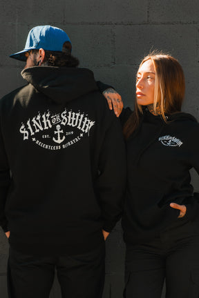 A man and woman wear Relentless Betrayal's Dark Waters SOS Hoodie by a gray brick wall; he shows off the back design, she faces him with her hand on her hip. This unisex hoodie elevates any look.