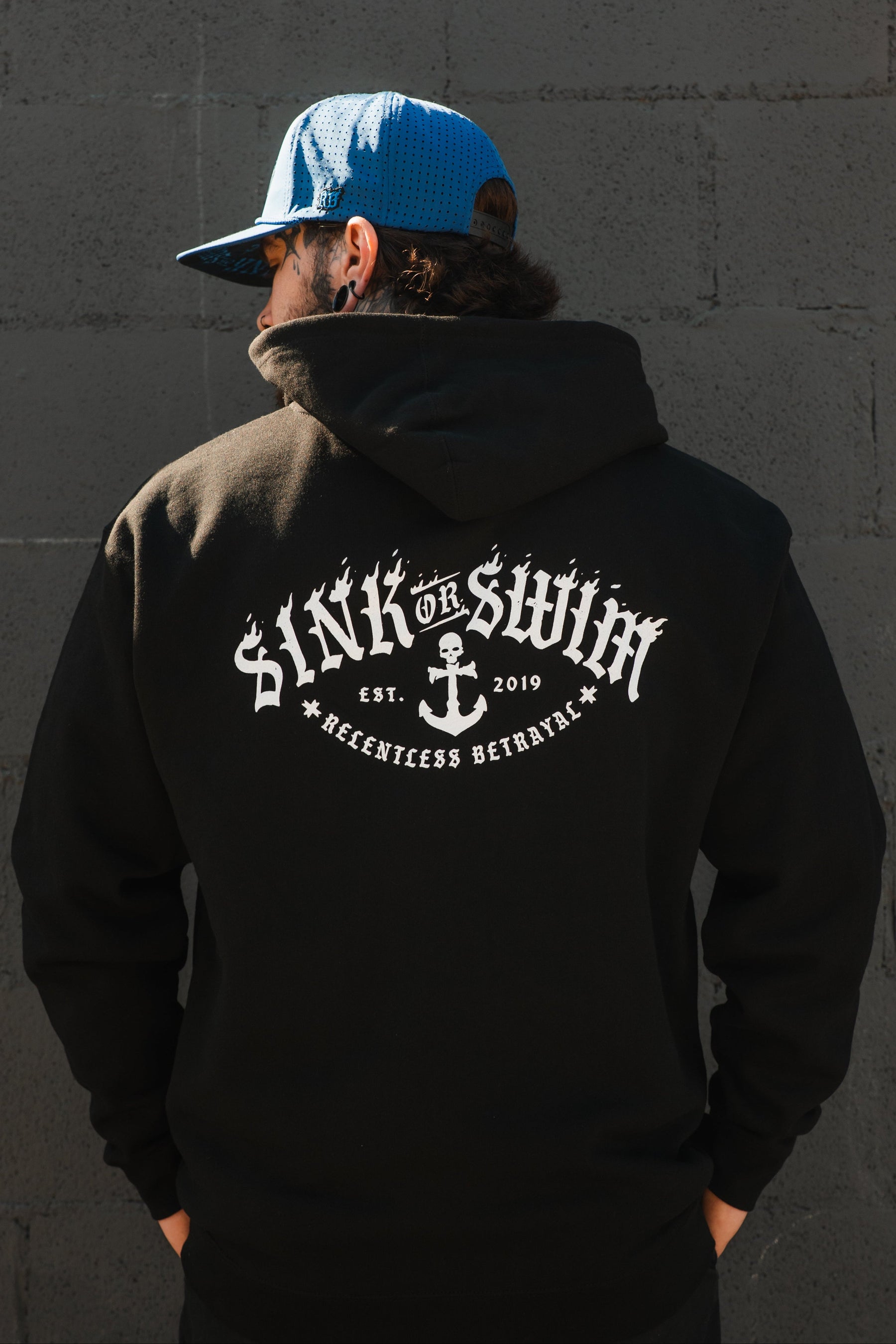 A person in a blue cap and black hoodie stands with their back to the camera. The unisex Relentless Betrayal Dark Waters SOS Hoodie features “SINK OR SWIM EST. 2019 RELENTLESS BETRAYAL” and an anchor graphic on the back.