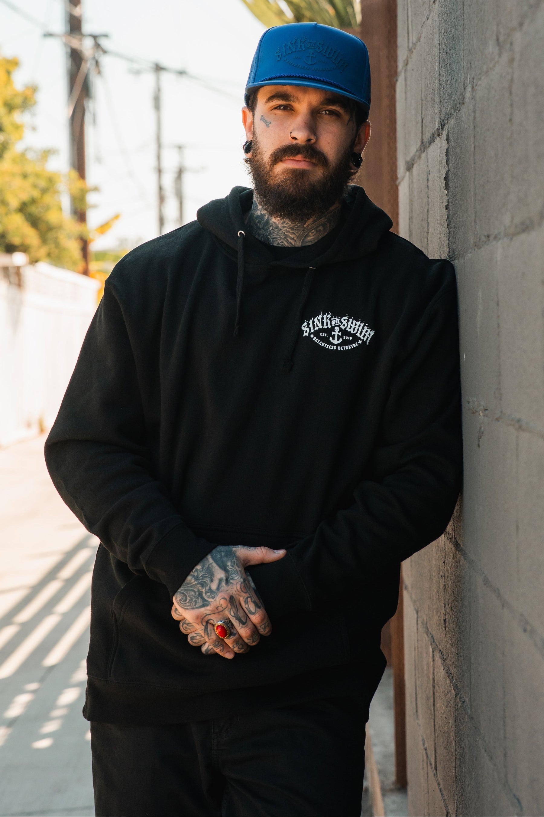 A bearded man with tattoos, wearing a blue cap and a black Relentless Betrayal Dark Waters SOS Hoodie, stands outdoors against a sunlit concrete wall, looking at the camera with his hands clasped.