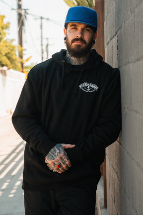 A bearded man with tattoos, wearing a blue cap and a black Relentless Betrayal Dark Waters SOS Hoodie, stands outdoors against a sunlit concrete wall, looking at the camera with his hands clasped.