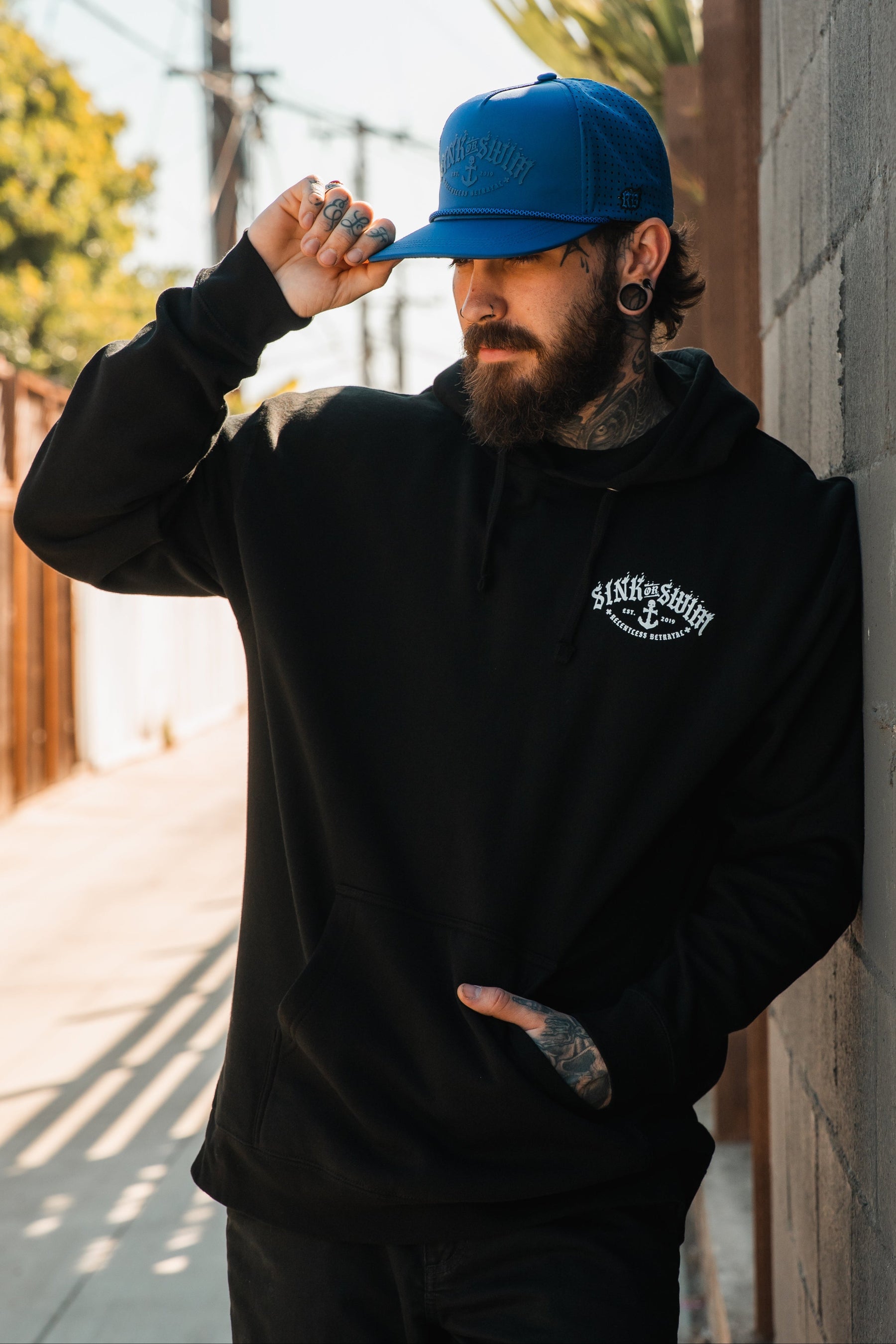 A tattooed bearded man, wearing a Relentless Betrayal Dark Waters SOS Hoodie and a bright blue cap, stands outdoors against a wall, holding his cap’s brim as sunlight casts shadows on the urban pathway nearby.