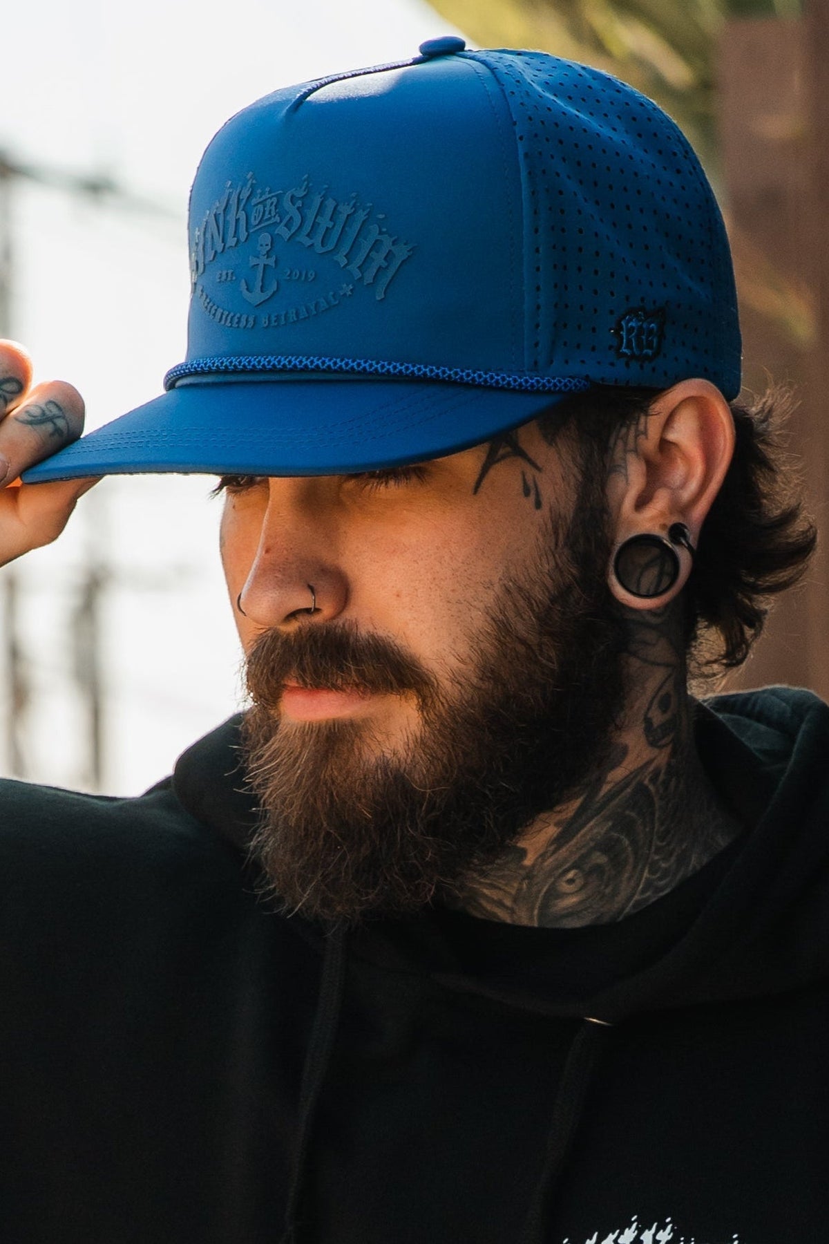 A bearded, tattooed person with stretched earlobes and a nose ring adjusts a Relentless Betrayal Dark Waters SOS Mute PVC Snapback while wearing a black hoodie outdoors in sunlight.