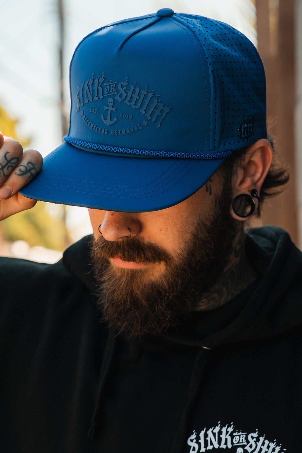 A tattooed, bearded person with piercings wears the Relentless Betrayal Dark Waters SOS Mute PVC Snapback and a black hoodie, embodying a pirate vibe. Outdoors, they look down while holding the cap’s brim.