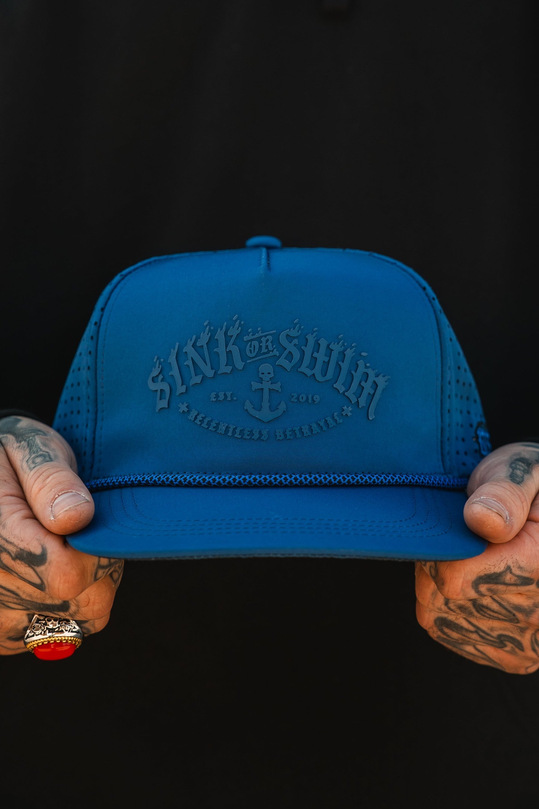 A tattooed hand with a ring holds the Relentless Betrayal Dark Waters SOS Mute PVC Snapback, featuring “Sink or Swim,” an anchor, and Est. 2019 on the blue patch—perfect for embracing a pirate vibe against a dark backdrop.