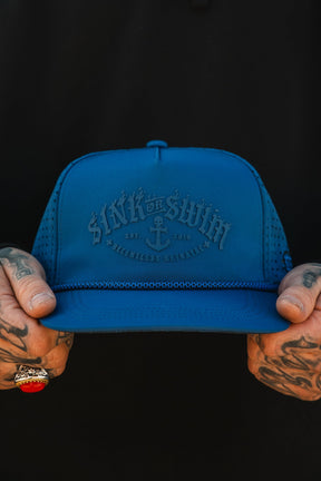 A tattooed hand with a ring holds the Relentless Betrayal Dark Waters SOS Mute PVC Snapback, featuring “Sink or Swim,” an anchor, and Est. 2019 on the blue patch—perfect for embracing a pirate vibe against a dark backdrop.