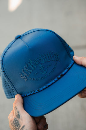 A person with tattooed fingers holds the Relentless Betrayal Dark Waters SOS Mute PVC Snapback, featuring "SINK OR SWIM" above an anchor and "RELENTLESS OBSESSION," capturing a bold, pirate-inspired vibe.