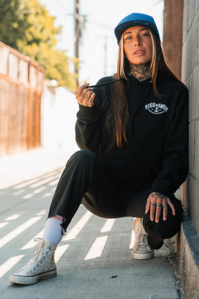 A tattooed woman in a Relentless Betrayal Dark Waters SOS Hoodie, black pants, and white high-top sneakers squats by a sunlit wall, wearing a blue cap and holding a cigarette.