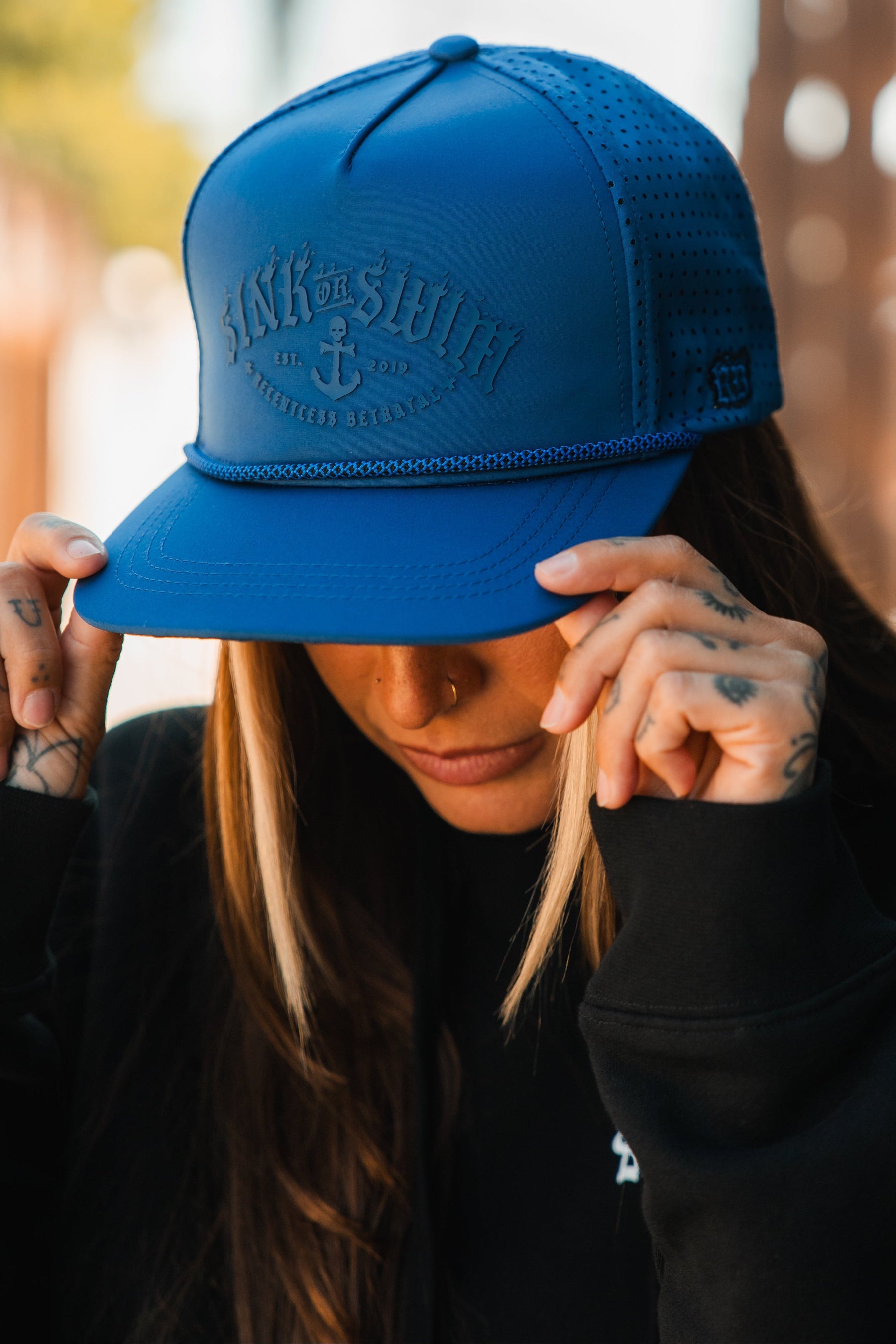A person with long hair and hand tattoos adjusts the brim of a bright blue Dark Waters SOS Mute PVC Snapback by Relentless Betrayal, its embossed text and anchor design evoking a distinctive pirate vibe.