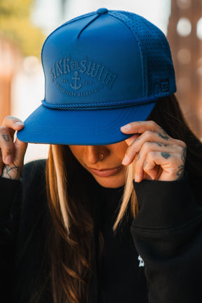 A person with long hair and hand tattoos adjusts the brim of a bright blue Dark Waters SOS Mute PVC Snapback by Relentless Betrayal, its embossed text and anchor design evoking a distinctive pirate vibe.