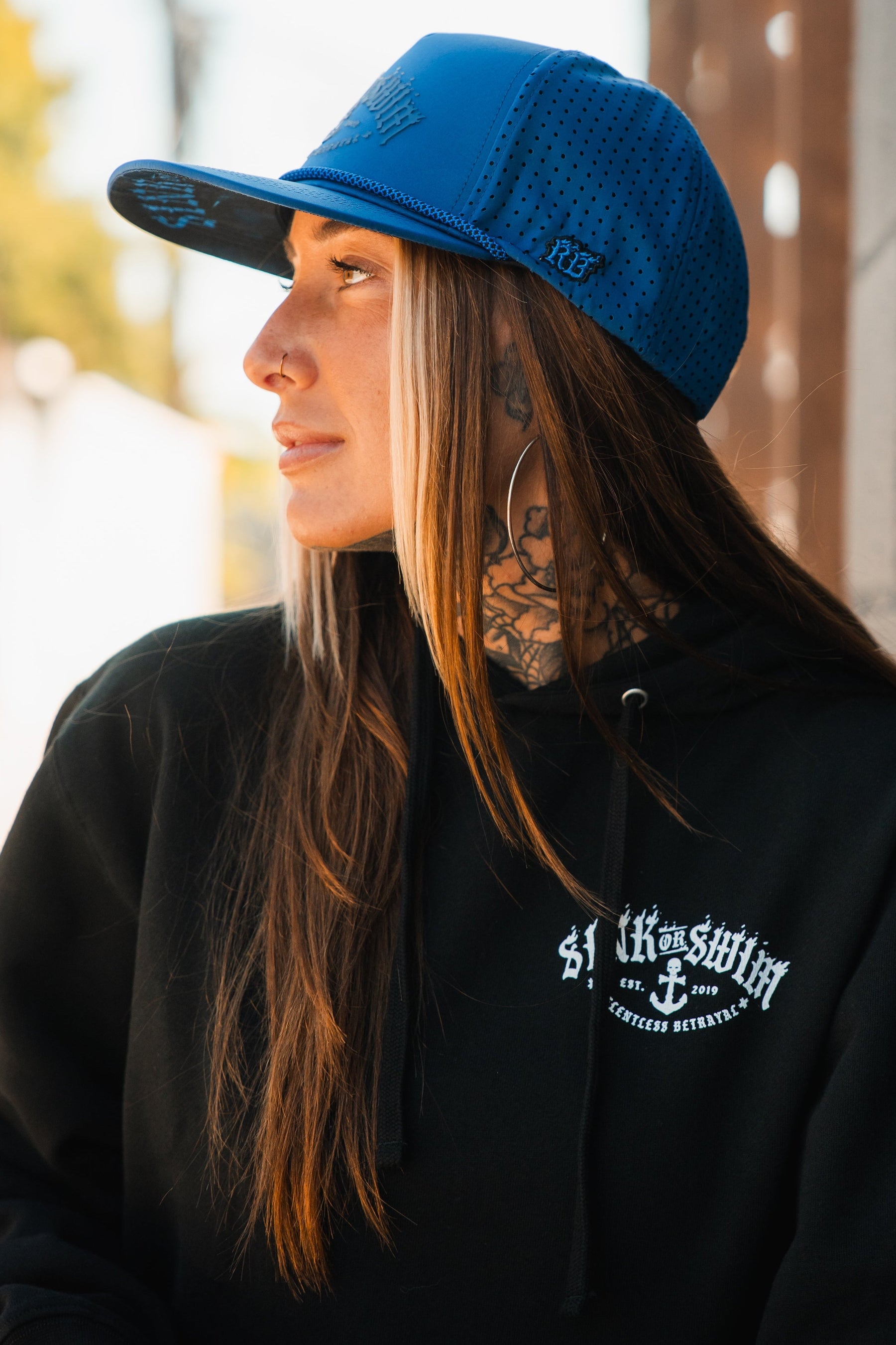 A woman with long brown hair, neck tattoos, and large hoop earrings wears a black hoodie and the Relentless Betrayal Dark Waters SOS Mute PVC Snapback in bright blue, looking to the side outdoors.
