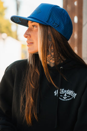 A woman with long brown hair, neck tattoos, and large hoop earrings wears a black hoodie and the Relentless Betrayal Dark Waters SOS Mute PVC Snapback in bright blue, looking to the side outdoors.
