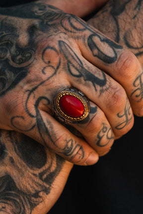 A close-up of tattooed hands with curled fingers, wearing the Burning Rose Ring by Relentless Betrayal—an artisanal statement piece featuring an oval red stone set in a detailed gold and silver band.