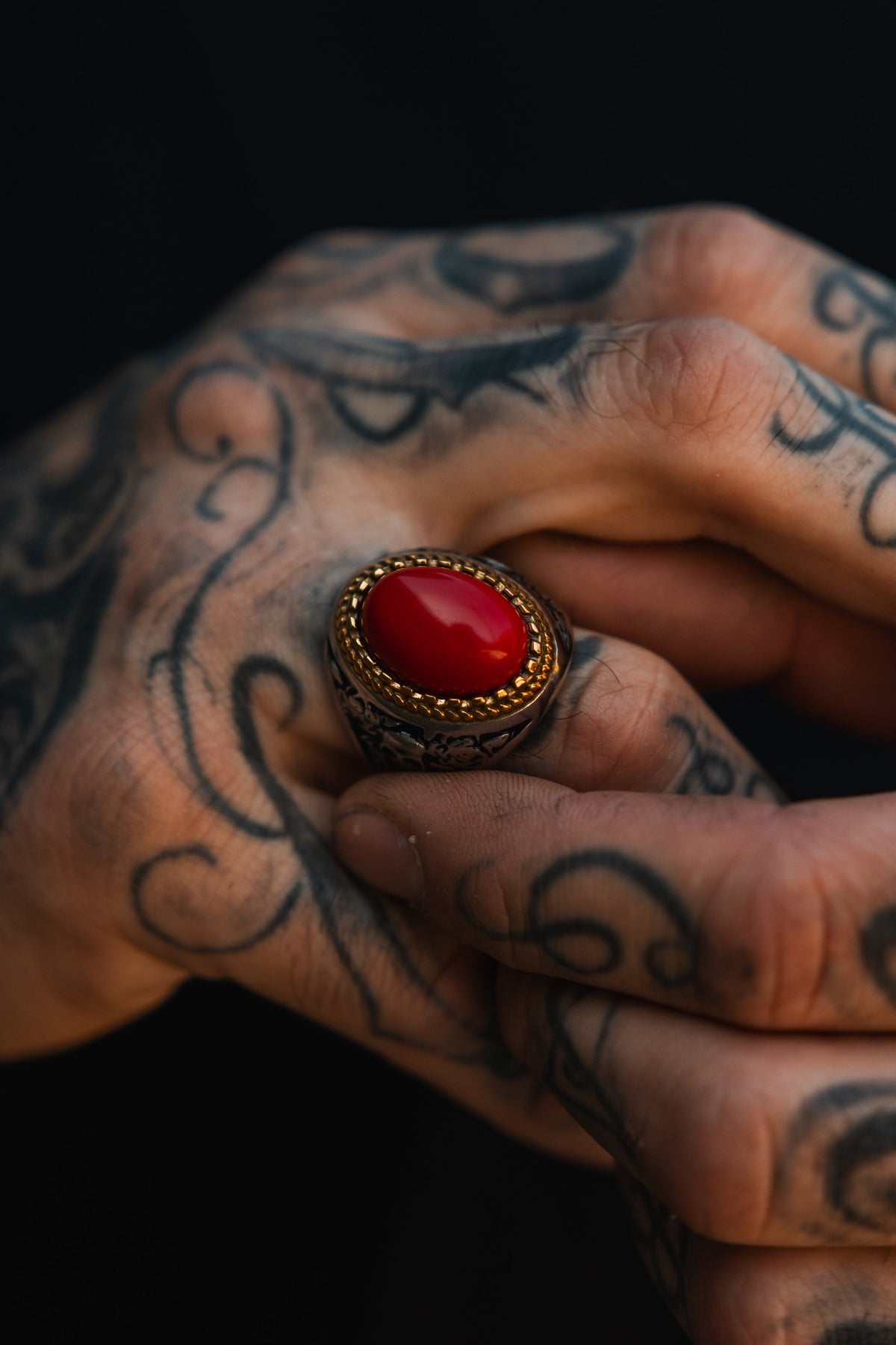 Tattooed hands hold the Burning Rose Ring by Relentless Betrayal, a handcrafted piece with a vivid red stone set in an intricately detailed metal band.