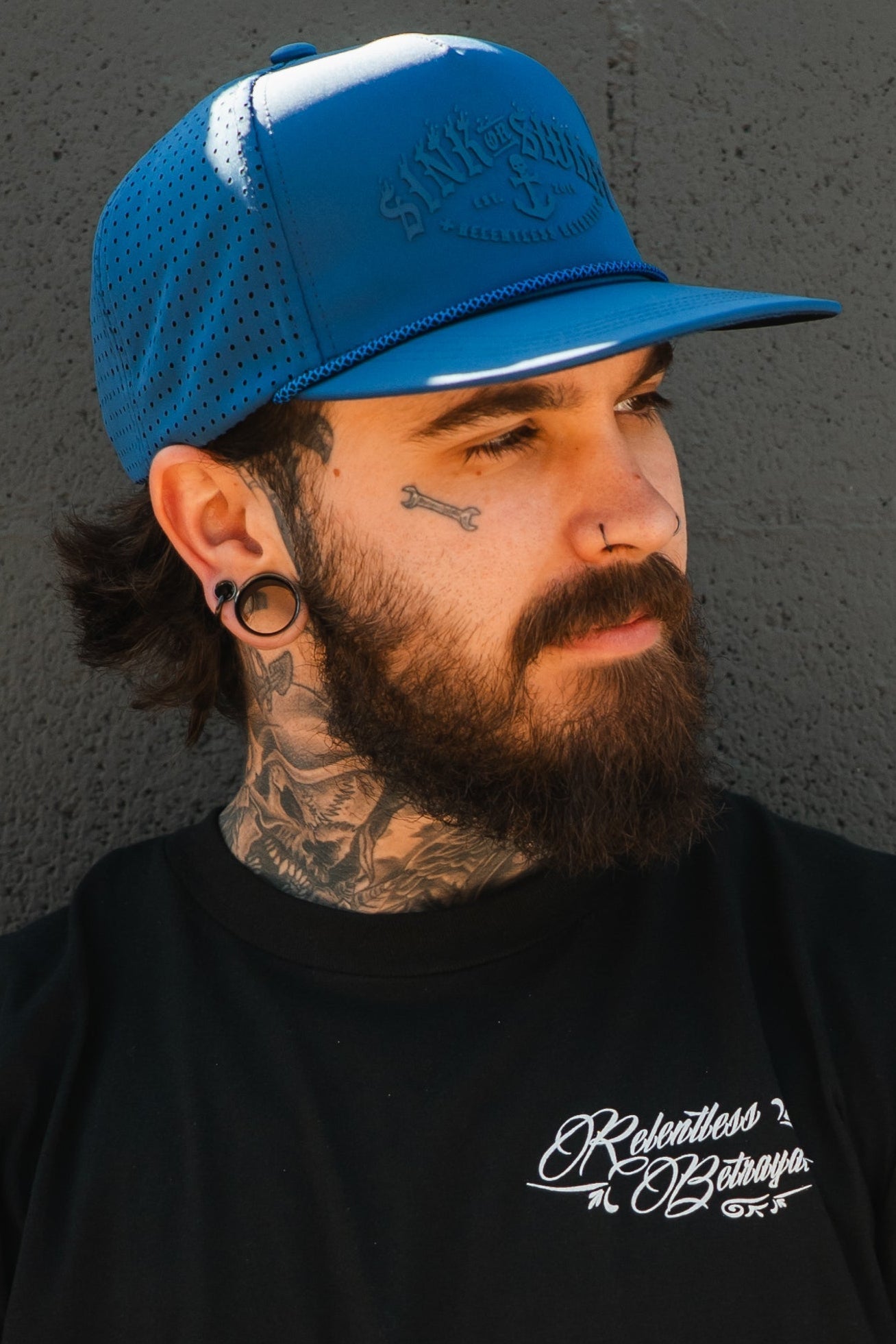 A bearded man with tattoos and stretched earlobes wears the Relentless Betrayal Dark Waters SOS Mute PVC Snapback and a black Relentless Betrayal shirt, standing against a textured gray wall and looking to the side.