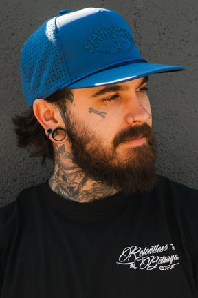 A bearded man with tattoos and stretched earlobes wears the Relentless Betrayal Dark Waters SOS Mute PVC Snapback and a black Relentless Betrayal shirt, standing against a textured gray wall and looking to the side.