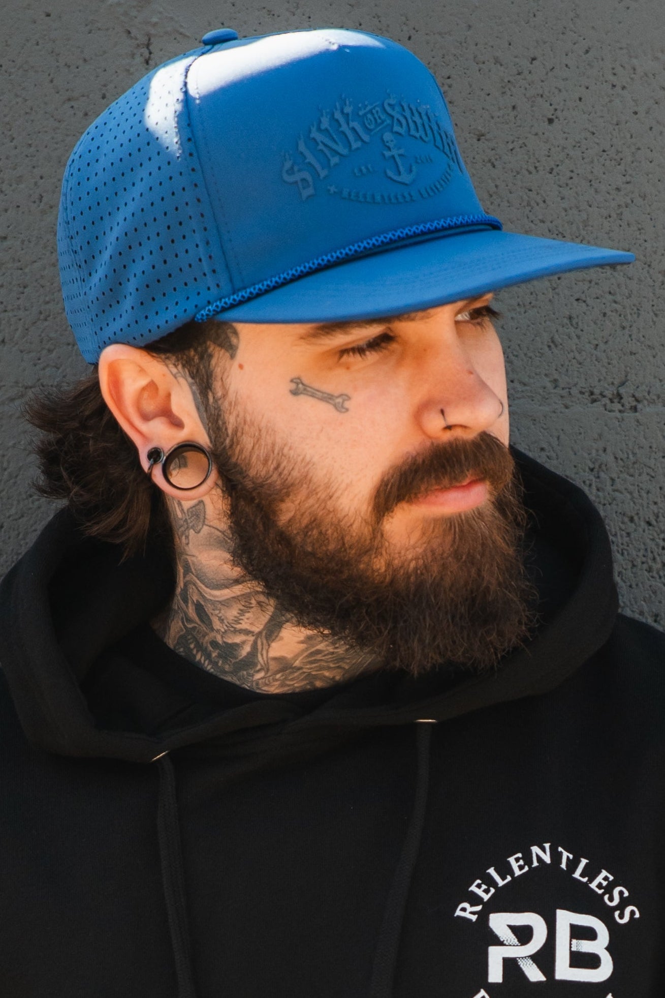 A man with a full beard, stretched earlobes, nose piercings, and tattoos wears the Relentless Betrayal Dark Waters SOS Mute PVC Snapback and a black hoodie, standing before a textured gray wall.