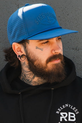 A man with a full beard, stretched earlobes, nose piercings, and tattoos wears the Relentless Betrayal Dark Waters SOS Mute PVC Snapback and a black hoodie, standing before a textured gray wall.