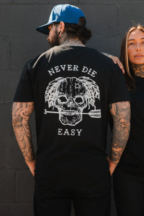 A tattooed man wears the Relentless Betrayal Never Die Easy Black Tee, made from fine jersey cotton and featuring a skull, feathers, an arrow, and "NEVER DIE EASY" on the back, standing next to a tattooed woman by a dark brick wall.