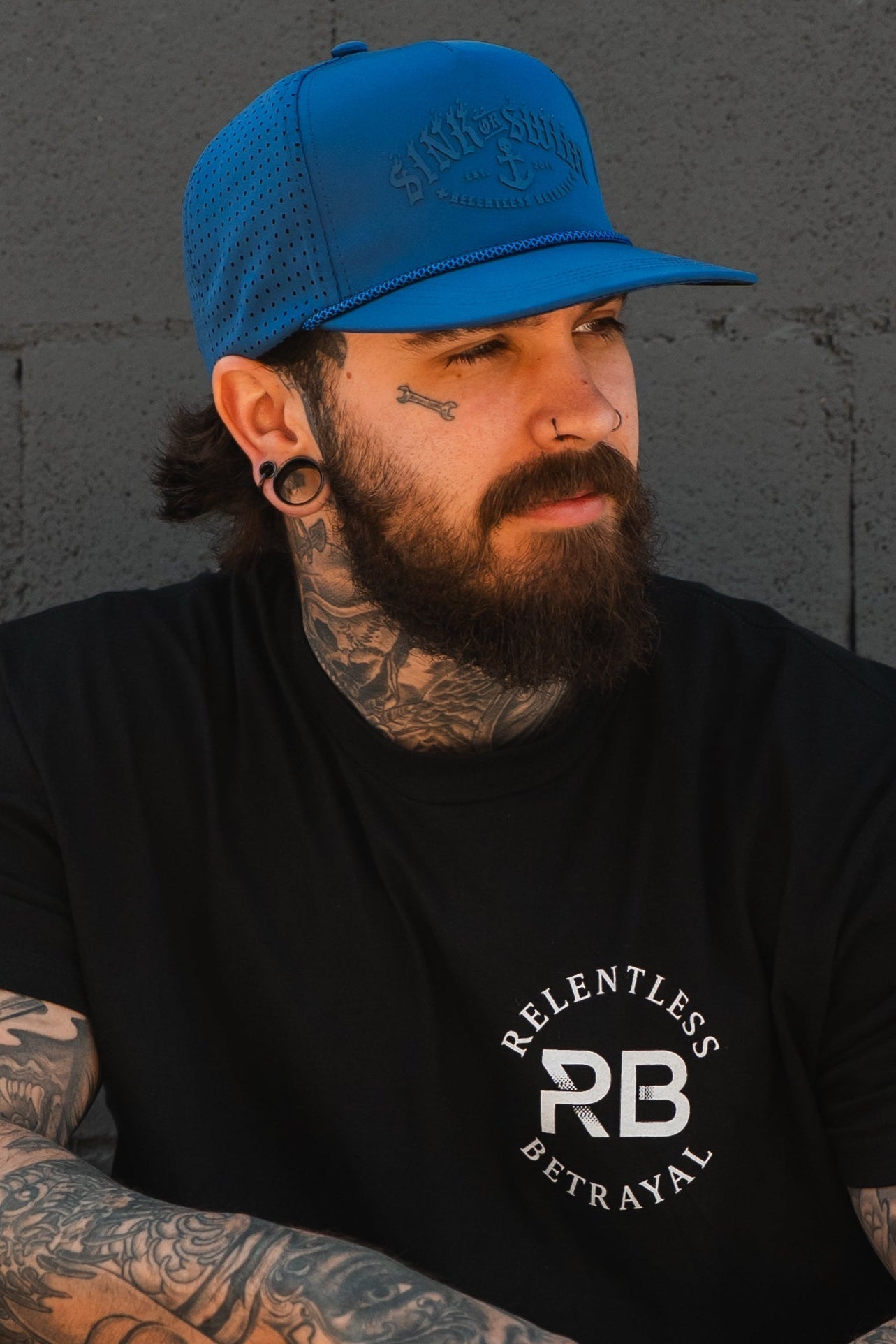 A bearded man with tattoos and piercings wears a Relentless Betrayal Dark Waters SOS Mute PVC Snapback and a black Relentless Betrayal t-shirt, standing against a gray concrete wall.