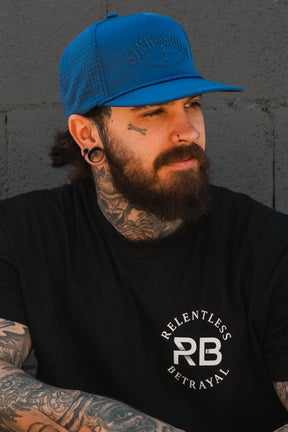A bearded man with tattoos and piercings wears a Relentless Betrayal Dark Waters SOS Mute PVC Snapback and a black Relentless Betrayal t-shirt, standing against a gray concrete wall.