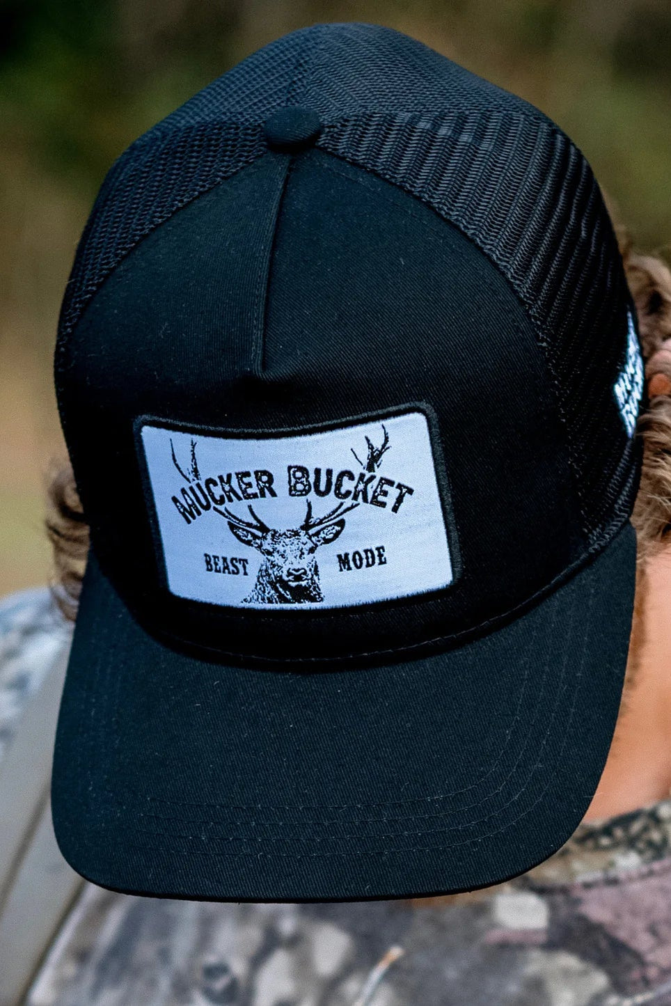 Wearing Mucker Buckets Beast Mode OG Trucker Bucket snapback, featuring a deer head design, and dressed in camo, they stand against a blurred green backdrop—ideal for any dedicated hunter or white tail deer enthusiast.