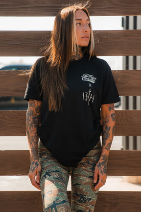 A woman with long brown hair and tattooed arms stands by a wooden fence, wearing the Tango Charlie Apparel "Lift Heavy. Pray Hard." black tee and camouflage leggings, gazing to the side with resilience.