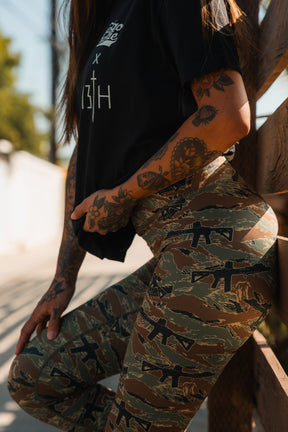 A woman with tattooed arms wears a Tango Charlie Apparel "Lift Heavy. Pray Hard." black tee and green camo rifle-patterned leggings, standing outdoors by a wooden fence in sunlight.