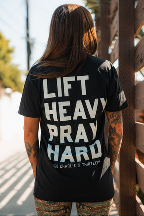 A tattooed individual stands outdoors in sunlight, wearing the Tango Charlie Apparel "Lift Heavy. Pray Hard." black tee—featuring bold white text—with wooden fencing in the background, embodying Faith Fitness.