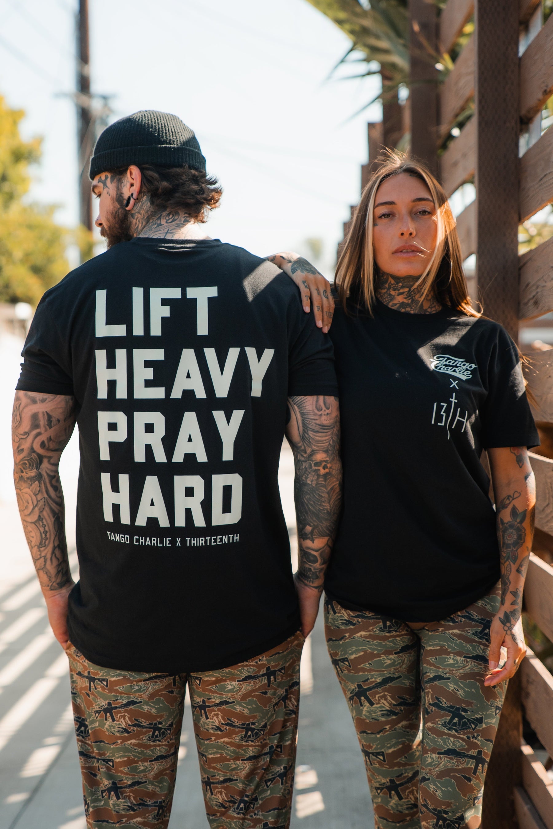 Two tattooed people in matching camo pants and black shirts stand outside. The person on the left wears the "Lift Heavy. Pray Hard." Black Tee by Tango Charlie Apparel, reflecting their Faith Fitness lifestyle; the other faces forward.