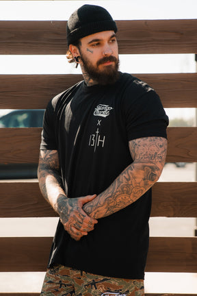 A bearded, tattooed man in the Lift Heavy. Pray Hard. - Black Tee by Tango Charlie Apparel stands before a wooden fence on a sunny day, hands clasped and gaze to the side—radiating spiritual fitness and quiet strength.