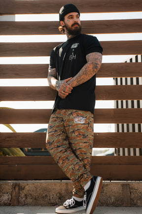 A bearded, tattooed man stands casually against a wooden fence in Tango Charlie Apparel’s "Lift Heavy. Pray Hard." black tee, camo pants, black skate shoes, and a beanie—arms crossed, he embodies true Faith Fitness spirit.