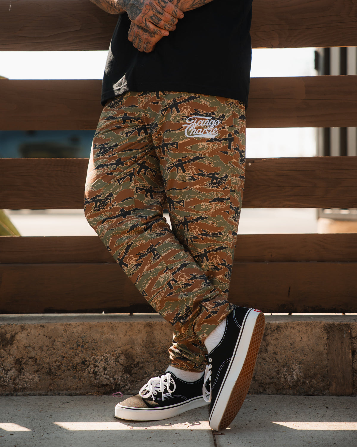 A person stands outdoors wearing Tango Charlie Apparel’s Men's Classic Premium Joggers | Snipes & Stripes—camouflage pants with Hando Master text—and black-and-white sneakers. Only the lower half shows, set against wooden fencing. Quick-dry fabric suits running.