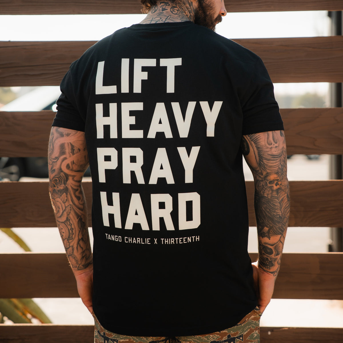 A tattooed man is seen from behind wearing the Lift Heavy. Pray Hard. - Black Tee by Tango Charlie Apparel, featuring bold white text and merging faith with fitness style.
