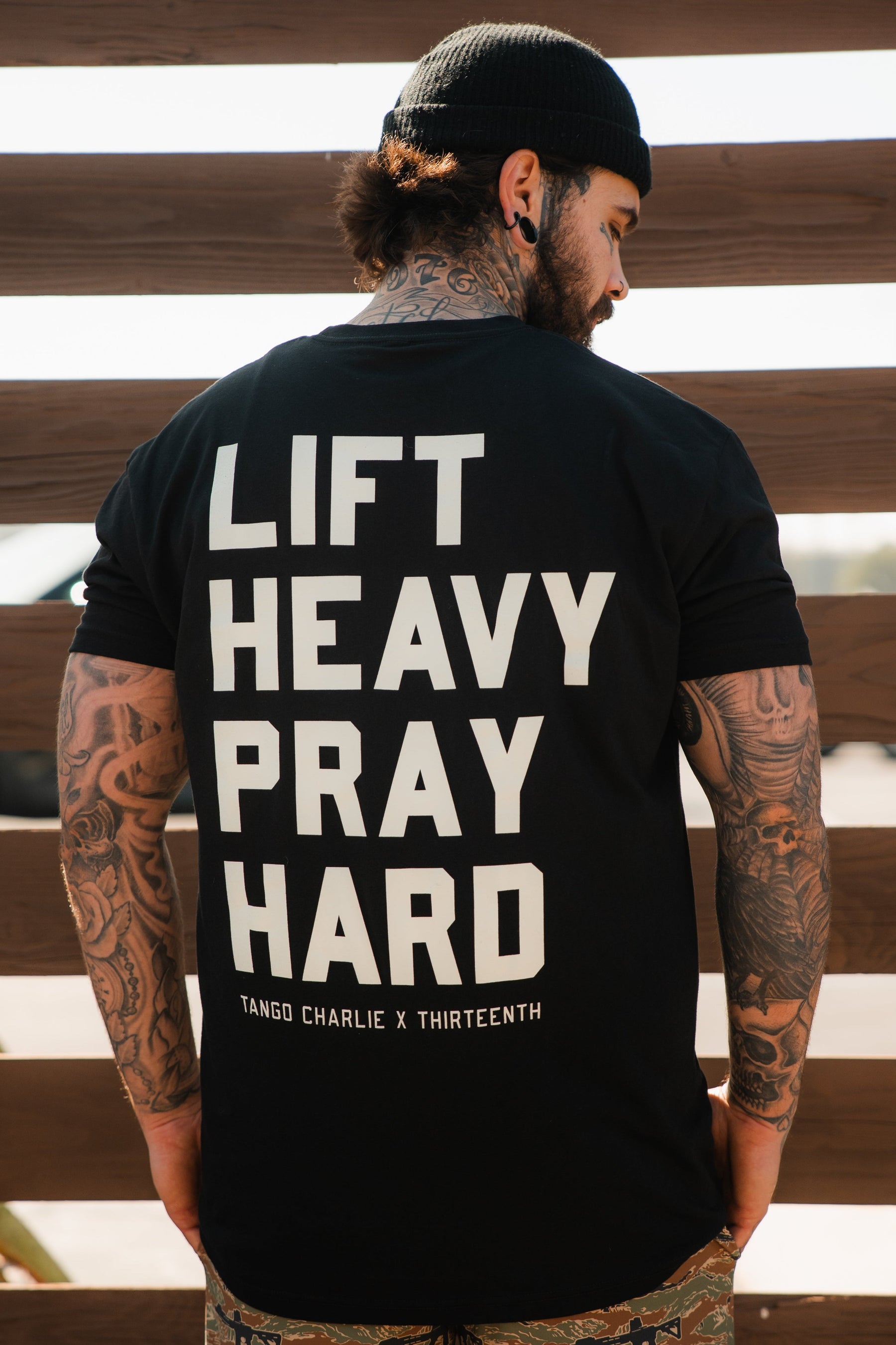 A tattooed man in a black beanie and a “Lift Heavy. Pray Hard.” Black Tee by Tango Charlie Apparel stands with his back to the camera before a wooden fence, capturing the spirit of spiritual fitness.