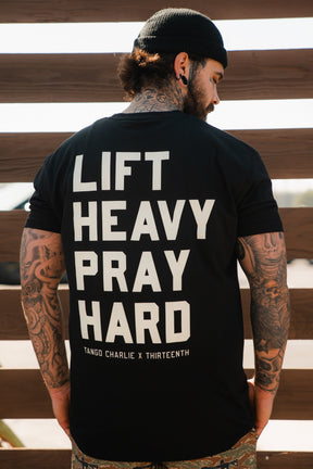 A tattooed man in a black beanie and a “Lift Heavy. Pray Hard.” Black Tee by Tango Charlie Apparel stands with his back to the camera before a wooden fence, capturing the spirit of spiritual fitness.