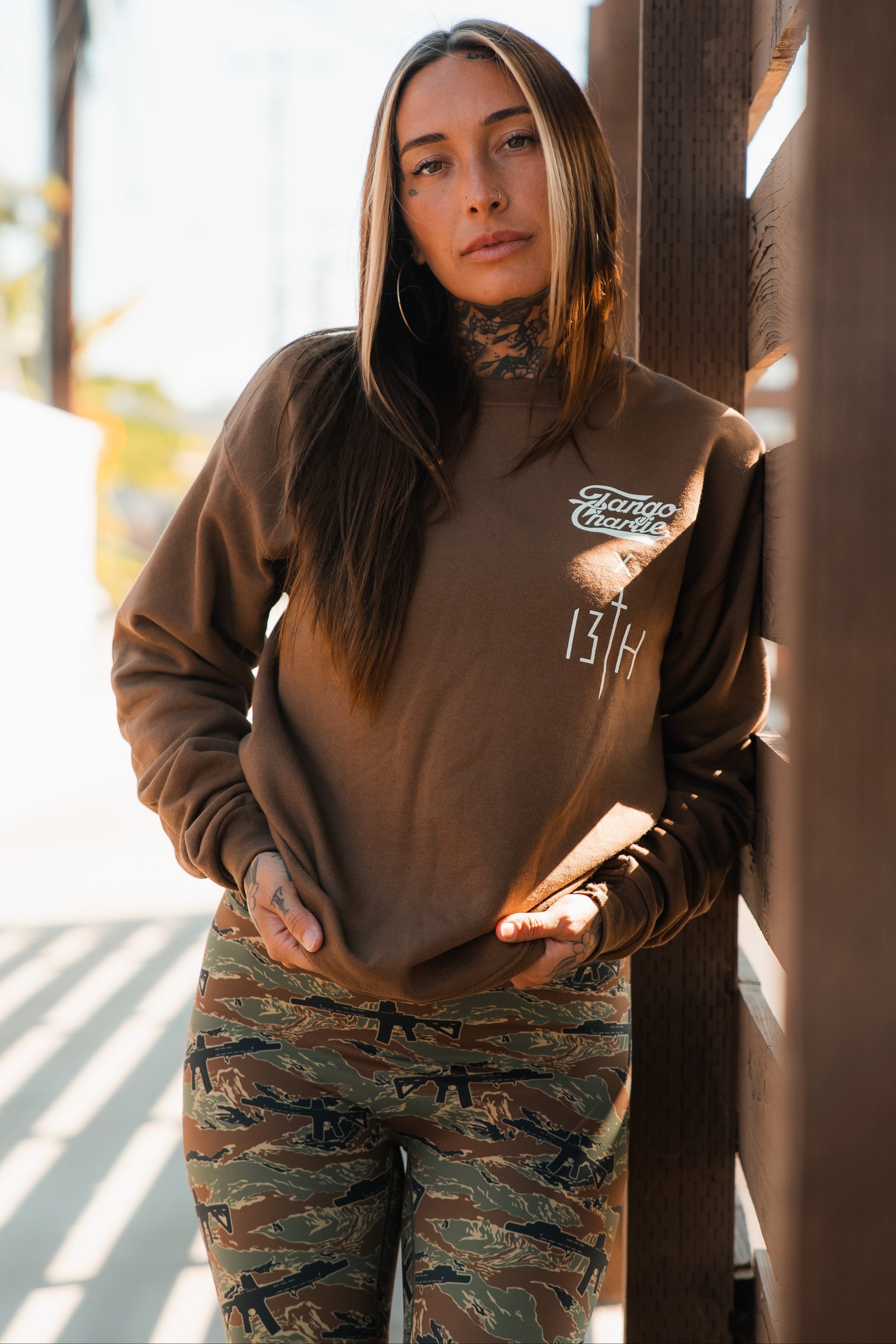 A woman with long hair and tattoos stands by a wooden fence in a brown Lift Heavy. Pray Hard. - Crewneck from Tango Charlie Apparel, embodying Christian fitness strength and spirit outdoors in camouflage pants.