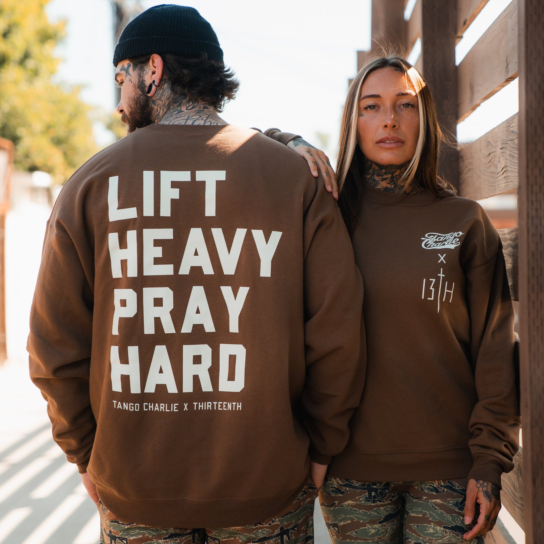 Two tattooed people in brown Tango Charlie Apparel crewnecks and camo pants stand outdoors. One sweatshirt reads "Lift Heavy. Pray Hard." on the back, while the other displays Tango Charlie x Thirteenth logos on the chest.