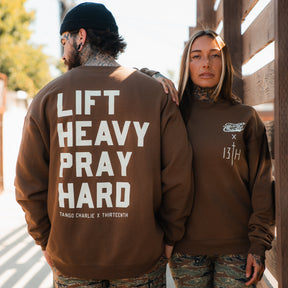 Two tattooed people in brown Tango Charlie Apparel crewnecks and camo pants stand outdoors. One sweatshirt reads "Lift Heavy. Pray Hard." on the back, while the other displays Tango Charlie x Thirteenth logos on the chest.